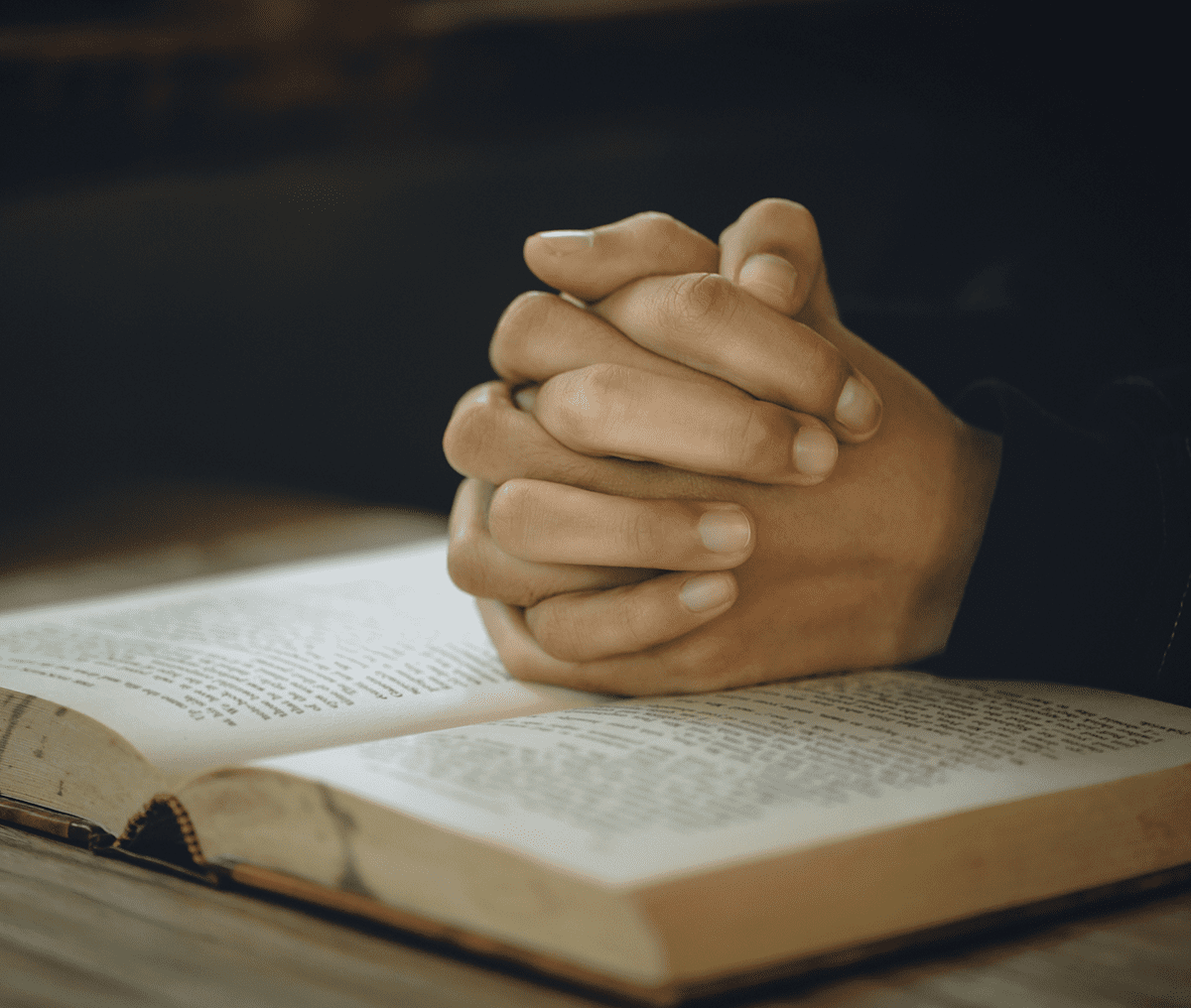 Hands clasped in prayer over an open book, likely a Bible.