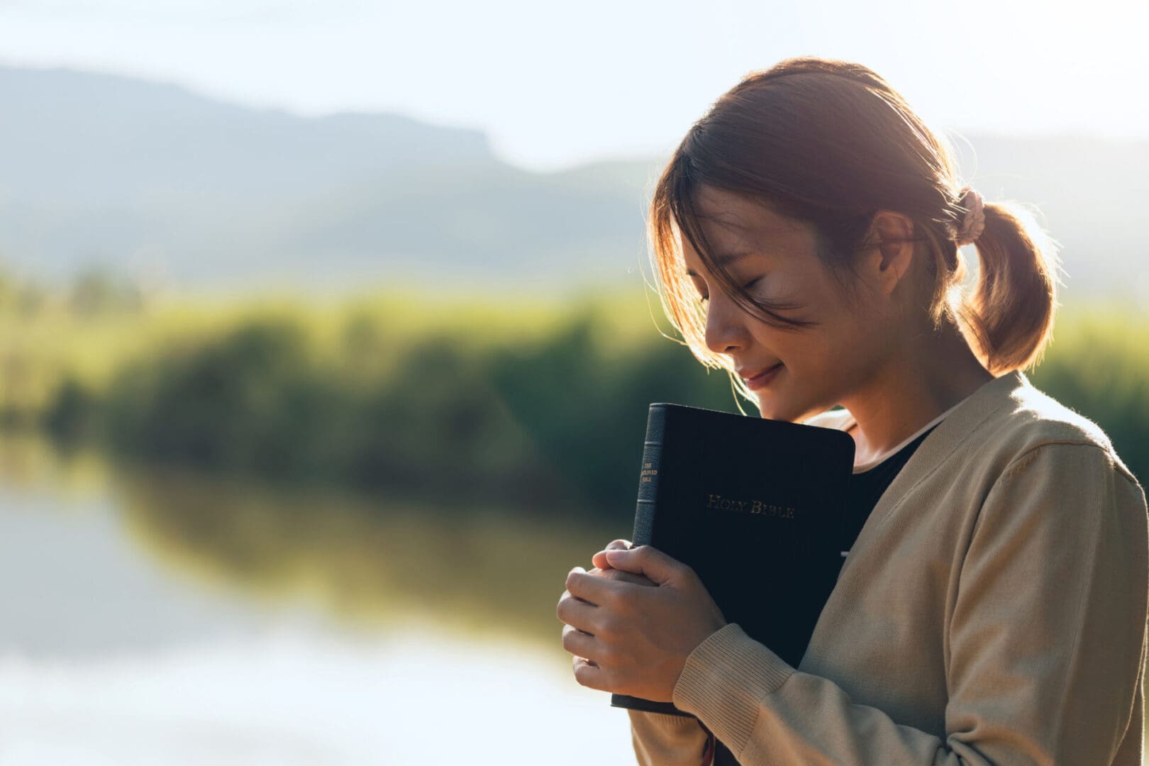 A woman holding a book, looking thoughtfully by a lake at sunset.