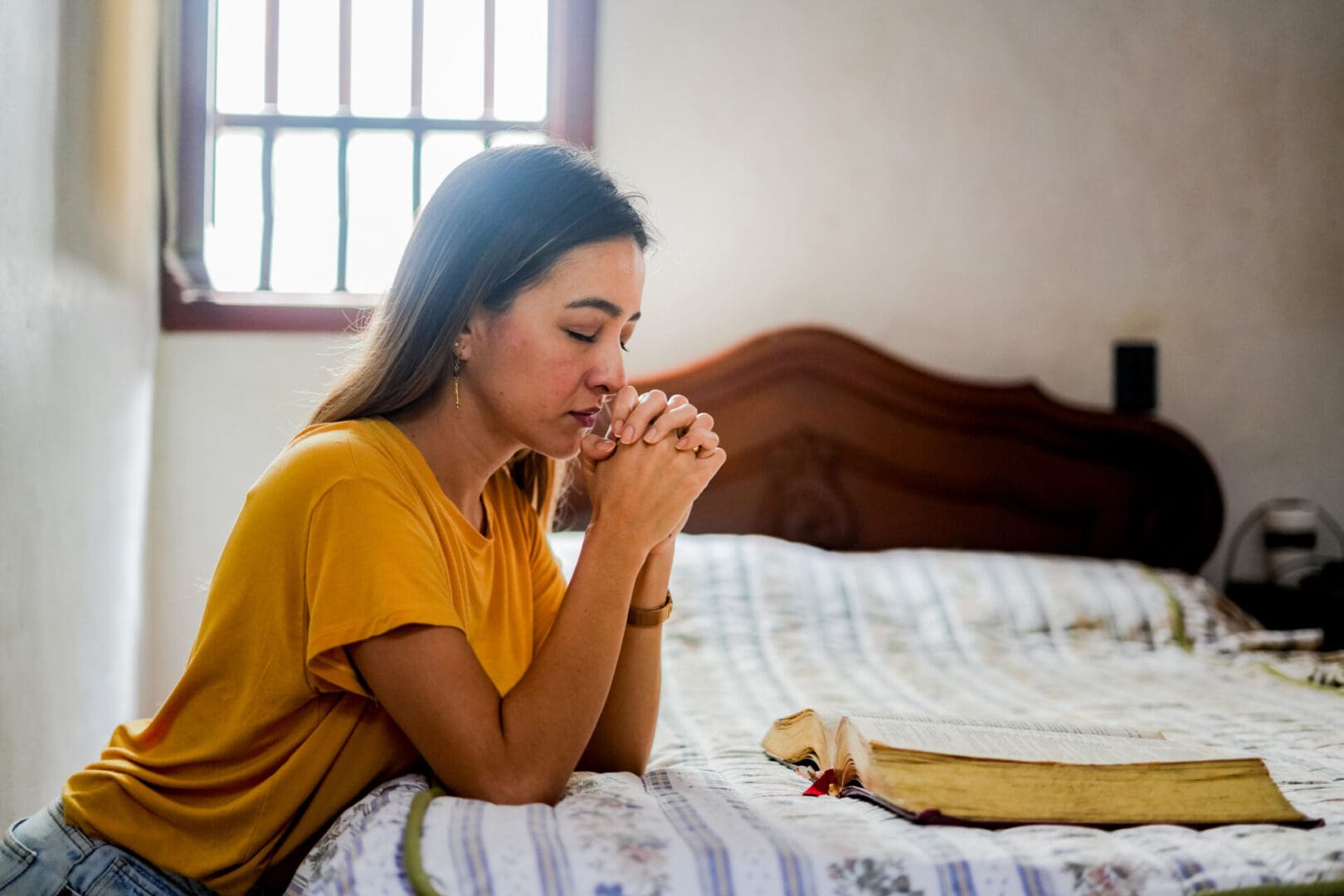 Woman praying with a Bible on the bed beside her.