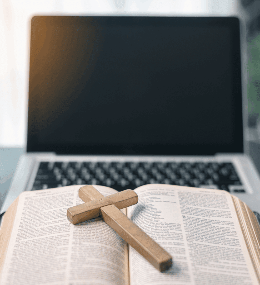 A wooden cross rests on an open Bible in front of a laptop.
