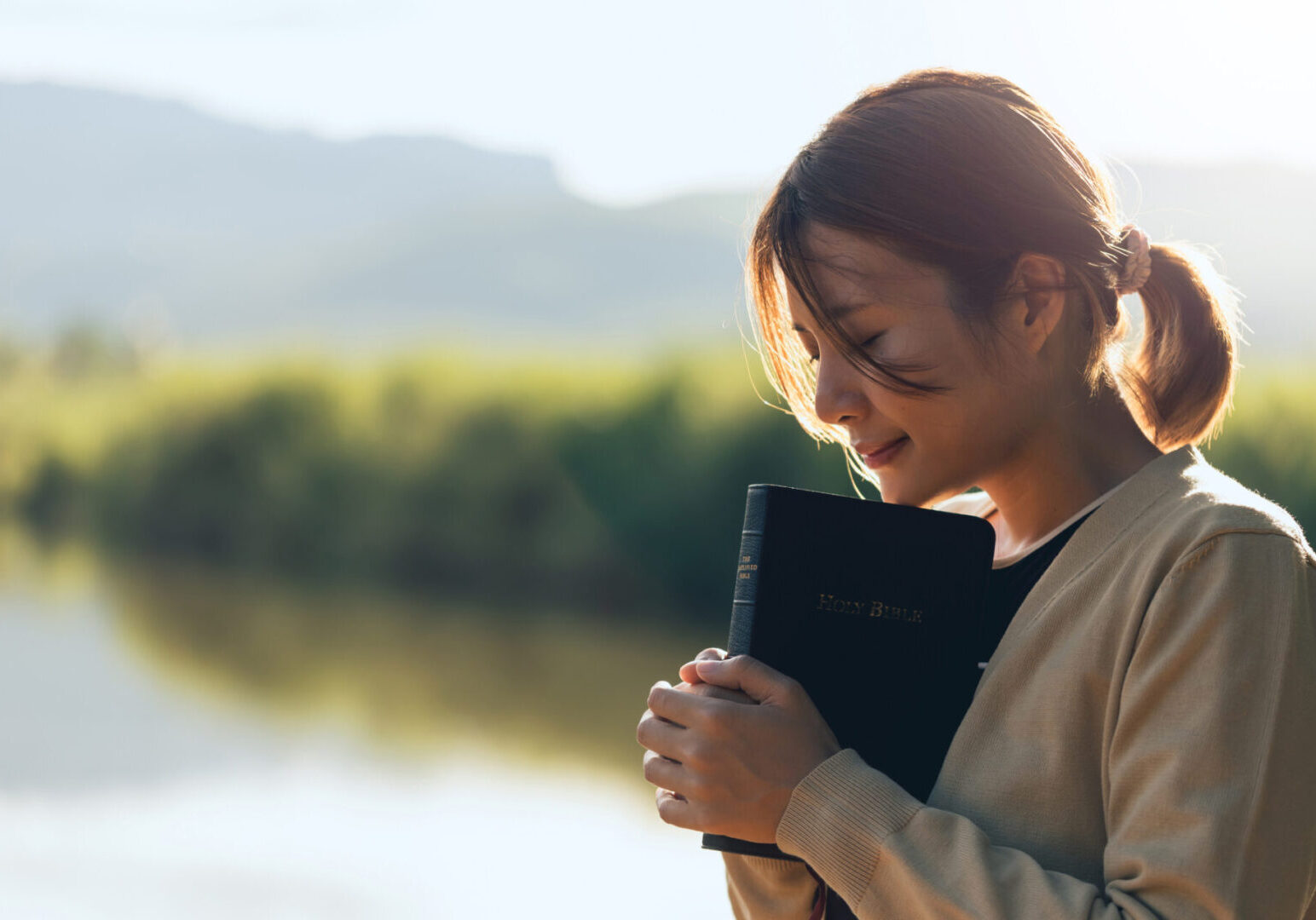 A woman holding a book, looking thoughtfully by a lake at sunset.