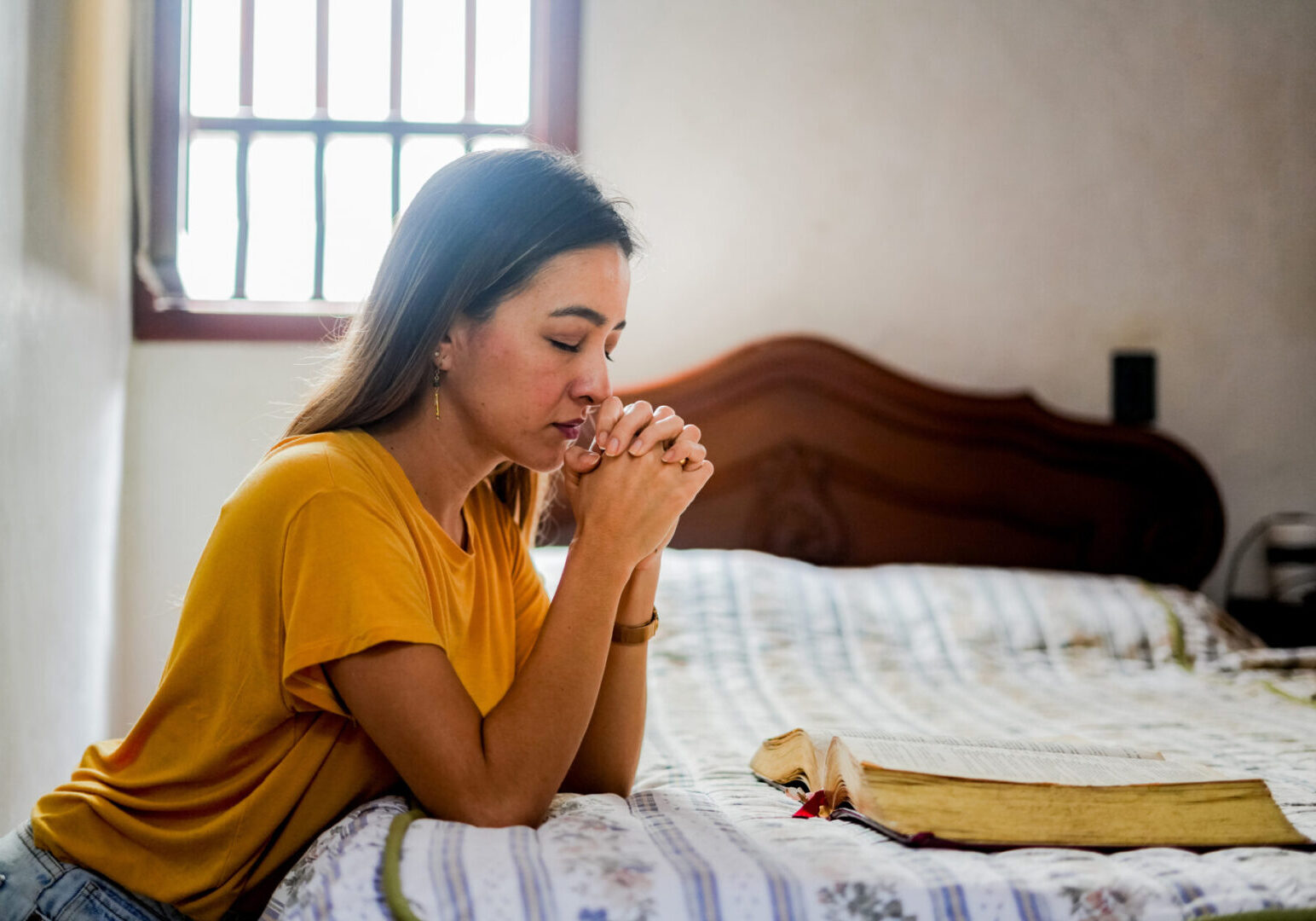 Woman praying with a Bible on the bed beside her.